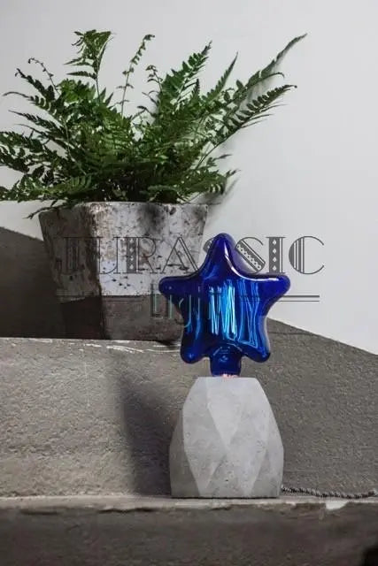 Ampoule étoile Star Blue en verre bleu sur un pied de lampe en béton, posée sur des marches près d'une fougère