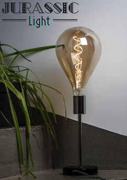 Ampoule LED en poire à filament spirale allumée sur un pied de lampe chromé, à côté d'une plante verte