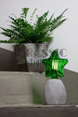 Ampoule étoile en verre vert allumée sur un pied de lampe en béton, posée sur des marches près d'une fougère