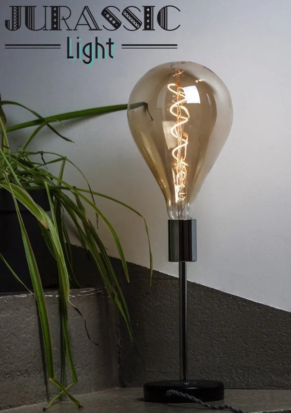 Ampoule LED en poire à filament spirale allumée sur un pied de lampe chromé, à côté d'une plante verte